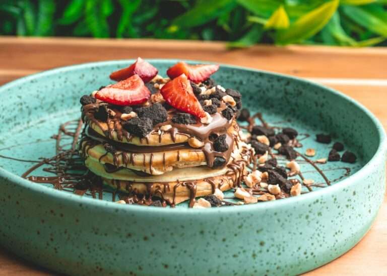 Der Food Fotograf - Ein Stapel Pfannkuchen mit Schokoladenüberzug, zerbröselten Keksen, gehackten Nüssen und geschnittenen Erdbeeren wird auf einem grünen Keramikteller serviert. Die Szene zeigt eine Holzoberfläche und grünes Laub im Hintergrund. Lecker Essen fotografieren.