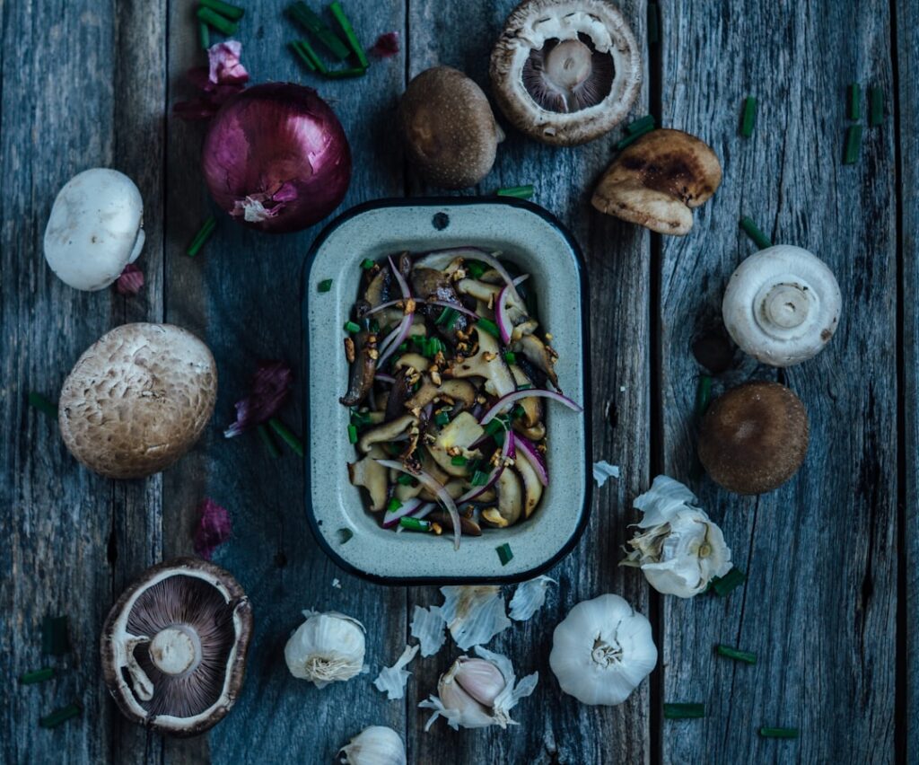 Food Fotograf Eine rechteckige Schüssel mit gebratenem Gemüse auf einer rustikalen Holzoberfläche, umgeben von verschiedenen Pilzen, Zwiebeln, Knoblauchzehen und gehackten grünen Kräutern. Essen fotografieren