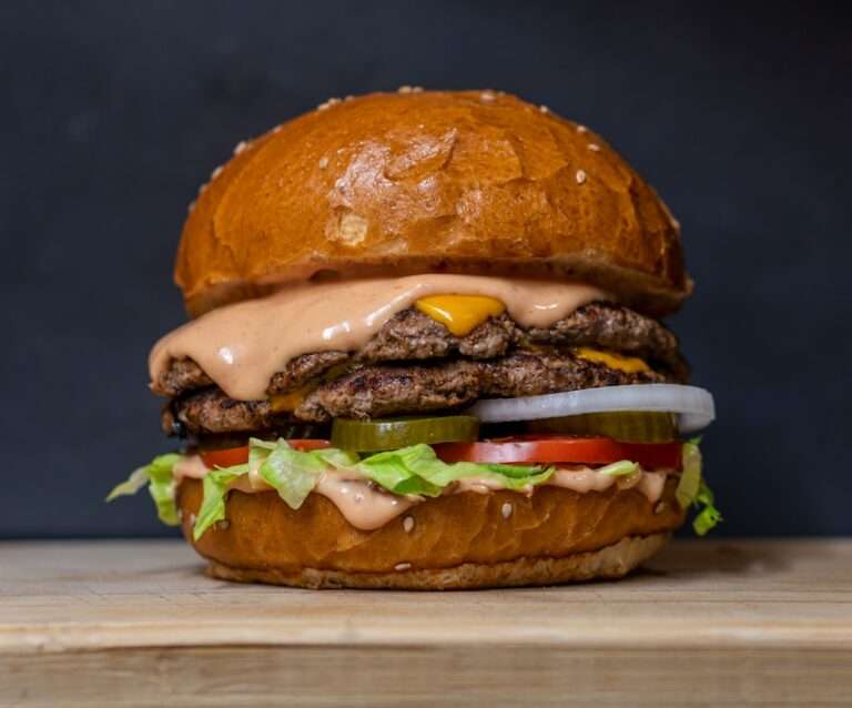 Der Food Fotograf - Eine Nahaufnahme eines Doppel-Cheeseburgers auf einer Holzoberfläche. Der Burger besteht aus zwei Rindfleisch-Patties mit geschmolzenem Käse, Gurkenscheiben, Zwiebeln, Salat, Tomaten und einer großzügigen Menge Soße, alles in einem Sesambrötchen. Lecker Essen fotografieren.