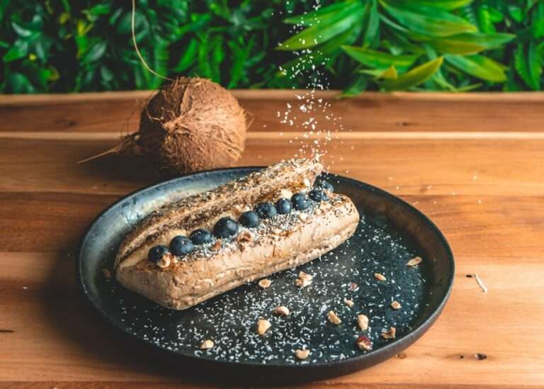 Der Food Fotograf - Ein Sandwich auf einem Teller, gefüllt mit Blaubeeren und bestreut mit Kokosraspeln. Das Sandwich liegt auf einem dunklen, runden Teller auf einer Holzoberfläche. Im Hintergrund sind eine ganze Kokosnuss und etwas grünes Laub zu sehen. Lecker Essen fotografieren.