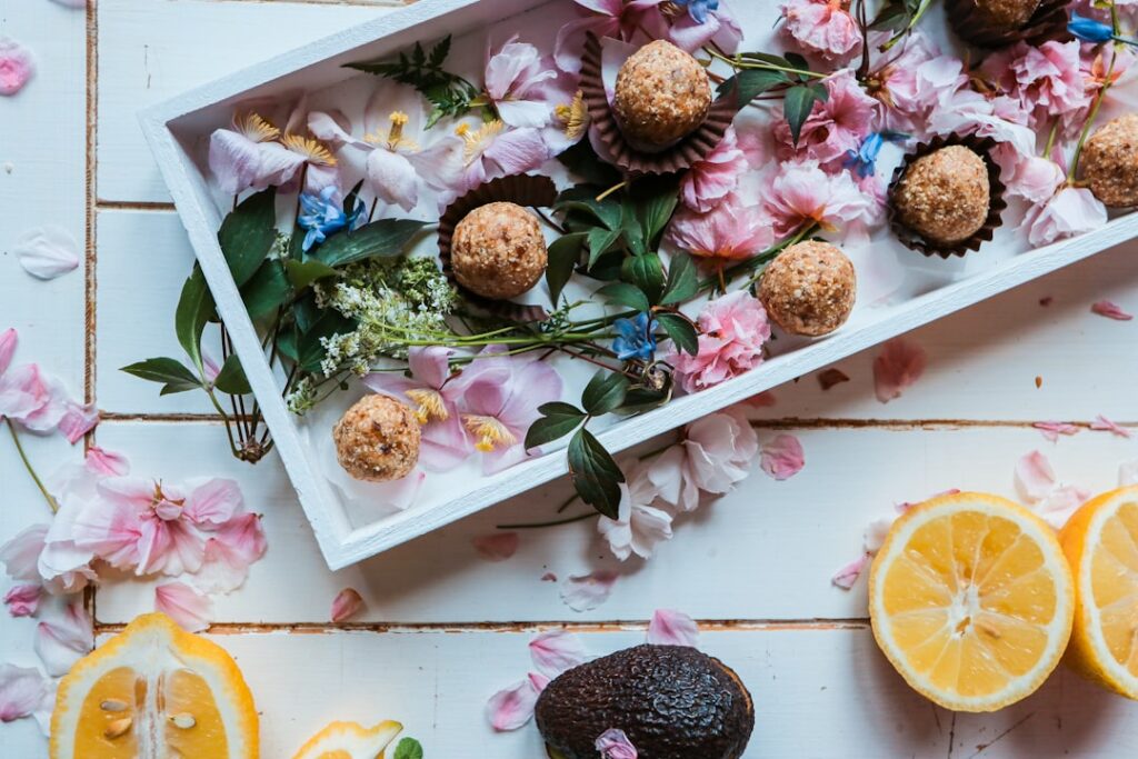 Food Fotograf Ein weißes, rechteckiges Tablett mit runden Süßigkeiten ist mit rosa Blütenblättern, grünen Blättern und anderen floralen Elementen geschmückt. Um das Tablett herum auf dem Tisch liegen halbierte Zitronen, eine Avocado und weitere verstreute rosa Blütenblätter und Blätter. Essen fotografieren