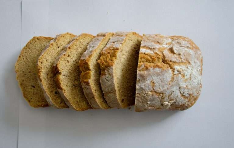 Der Food Fotograf - Ein in mehrere Stücke geschnittener Laib Brot liegt auf einer weißen Fläche. Das Brot scheint außen knusprig und grobkörnig zu sein, innen ist es weich und leicht krümelig. Die Scheiben sind ungleichmäßig geschnitten. Lecker Essen fotografieren.