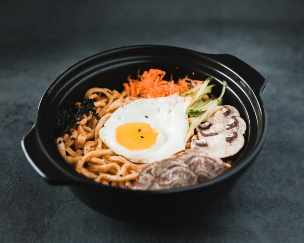Food Fotograf Eine schwarze Schüssel gefüllt mit Ramennudeln, garniert mit einem Spiegelei, geschnittenen Champignons, geraspelten Karotten und grünem Gemüse auf dunklem Hintergrund. Essen fotografieren