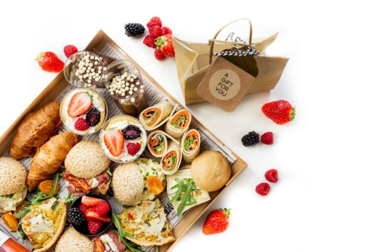 Food Fotograf Ein Tablett mit verschiedenen Lebensmitteln, darunter Croissants, Brötchen, Sandwiches, Obst und Desserts. Neben dem Tablett steht eine Geschenktüte mit einem Etikett mit der Aufschrift „EIN GESCHENK FÜR DICH“. Frische Erdbeeren, Brombeeren und Himbeeren sind überall verteilt. Essen fotografieren