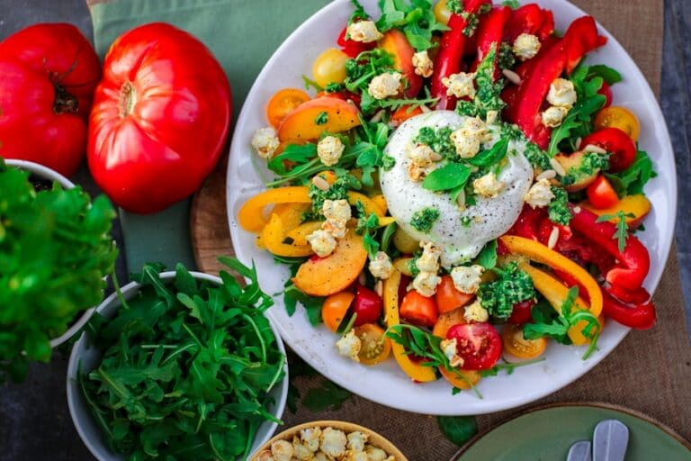 Food Fotograf Ein bunter Salat auf einem weißen Teller besteht aus Burrata-Käse, Rucola, roten und gelben Paprikaschoten, Kirschtomaten und Popcorn. Neben dem Teller stehen zwei Tomaten und eine Schüssel Rucola. Eine grüne Serviette und Besteck sind teilweise sichtbar. Essen fotografieren