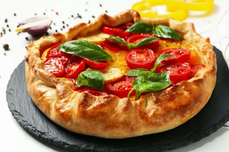 Food Fotograf Hannover - Auf einem schwarzen Schieferteller wird eine goldbraune, rustikale Torte mit Tomatenscheiben und frischen Basilikumblättern serviert. Im Hintergrund sind Pfefferkörner, ein Stück rote Zwiebel und gelbe Paprikascheiben verstreut. Food Fotografie Hannover.