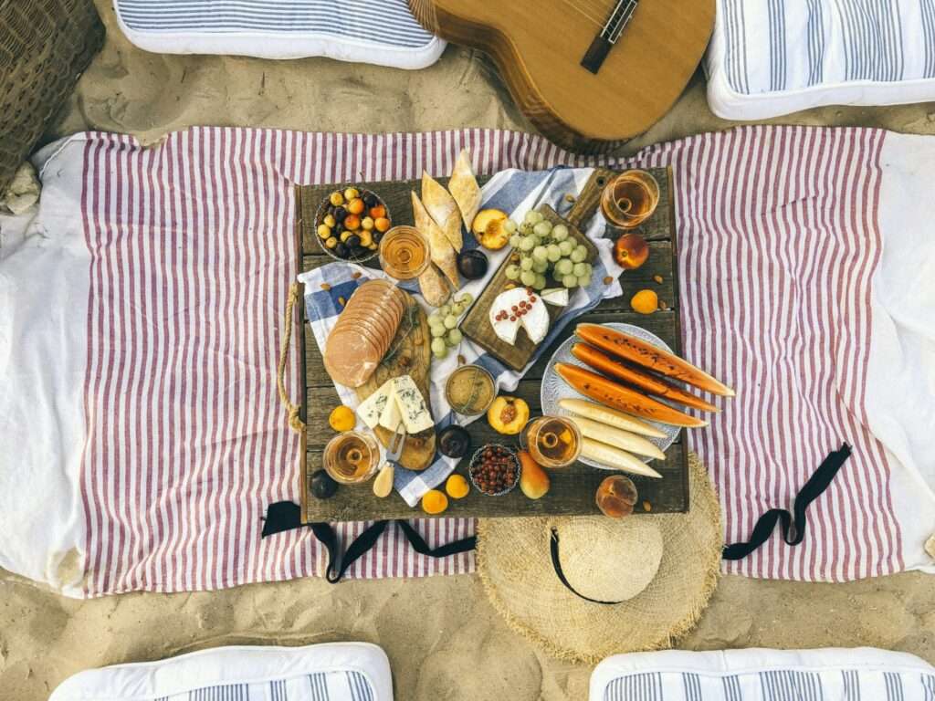 Food Fotograf Hannover - Ein Picknick-Setup im Sand umfasst ein Holztablett mit verschiedenen Käsesorten, Brot, Trauben, Pfirsichen, Wassermelonenscheiben und Nüssen. Um das Tablett herum liegen gestreifte Decken, Kissen, ein Hut und eine Gitarre. Auf dem Tablett stehen außerdem vier Getränke. Food Fotografie Hannover.
