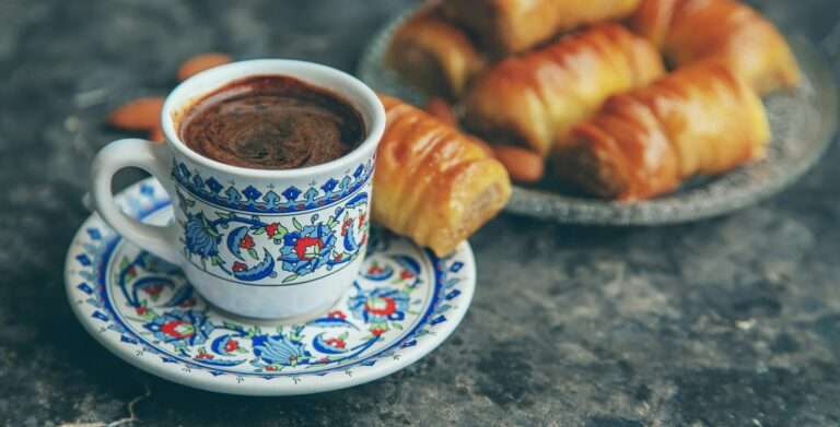Food Fotograf Hannover - Eine dekorierte Tasse und Untertasse mit Kaffee stehen auf einer Marmoroberfläche. Im Hintergrund ist ein Teller mit mehreren Croissants zu sehen. Die Tasse und Untertasse, die diese entzückende Lebensmittelfotografie einfangen, weisen komplizierte blaue, rote und weiße Blumenmuster auf. Food Fotografie Hannover.
