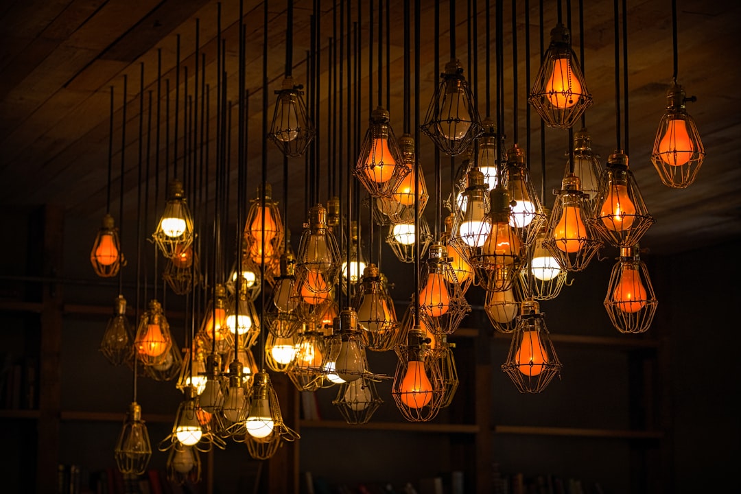 Der Food Fotograf - Eine Deckeninstallation aus Glühbirnen im Vintage-Stil in Drahtkäfigen, die abwechselnd beleuchtet werden und warmes orangefarbenes und gelbes Licht abgeben. Die Glühbirnen hängen an langen Kabeln und erzeugen ein komplexes und kunstvolles Lichtarrangement vor einem hölzernen Deckenhintergrund. Lecker Essen fotografieren.