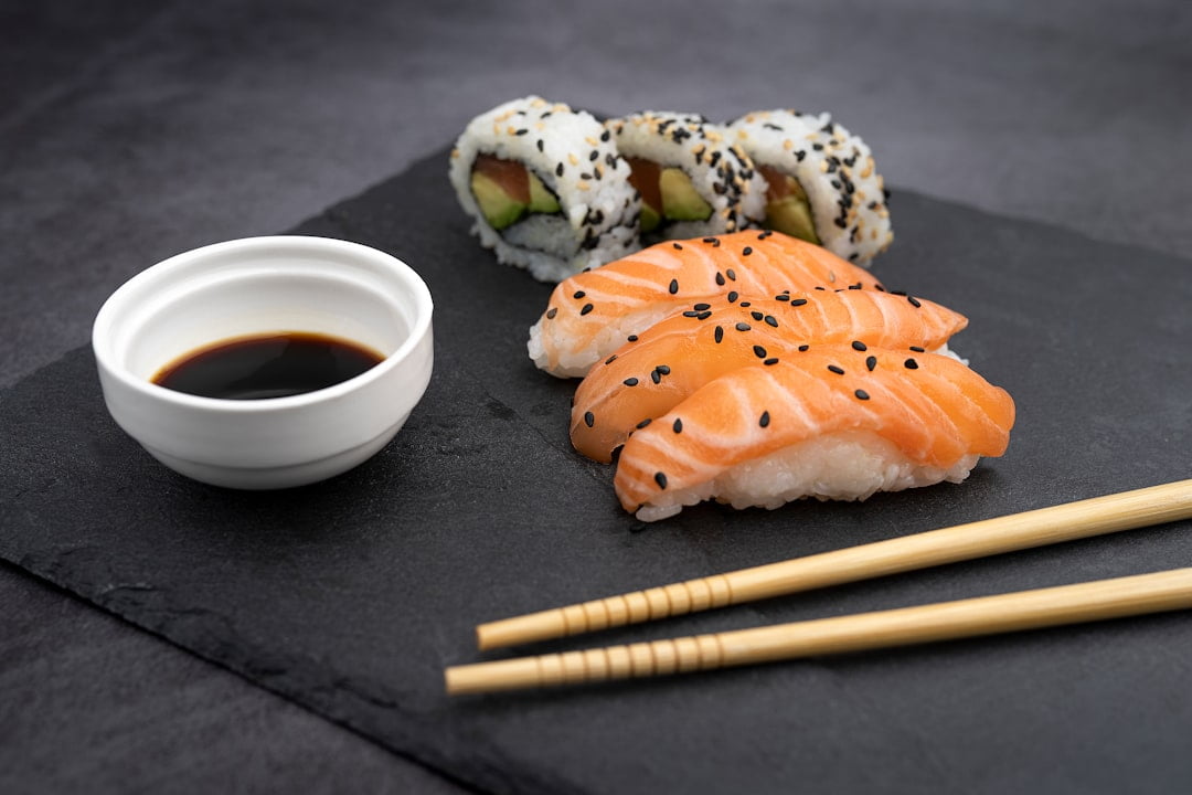 Der Food Fotograf - Ein Teller mit Sushi wird ausgestellt. Im Vordergrund sind Lachs-Nigiri mit schwarzem Sesam zu sehen. Im Hintergrund sind Uramaki-Sushi-Rollen mit Avocado und Fisch zu sehen. Neben dem Sushi stehen eine kleine Schüssel Sojasauce und ein Paar Essstäbchen. Lecker Essen fotografieren.