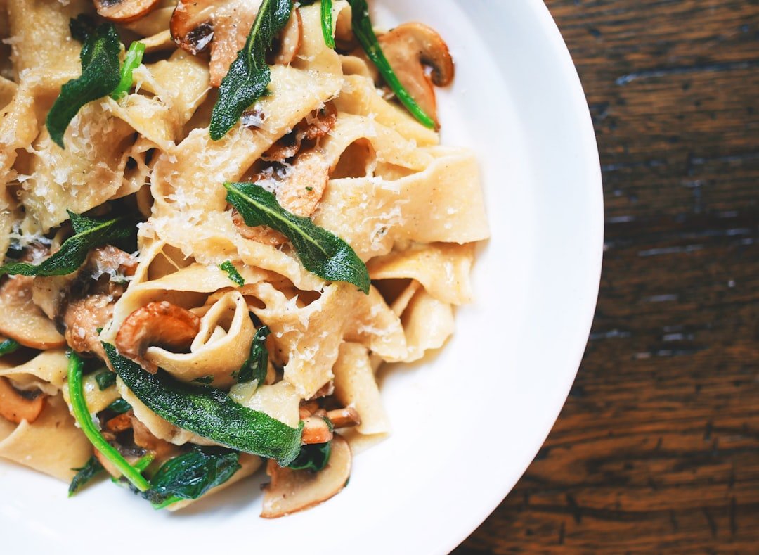 Der Food Fotograf - Ein weißer Teller mit Pappardelle-Nudeln ist mit sautierten Pilzen, frischem grünem Gemüse und geriebenem Käse garniert. Der Teller steht auf einer Holzoberfläche. Lecker Essen fotografieren.