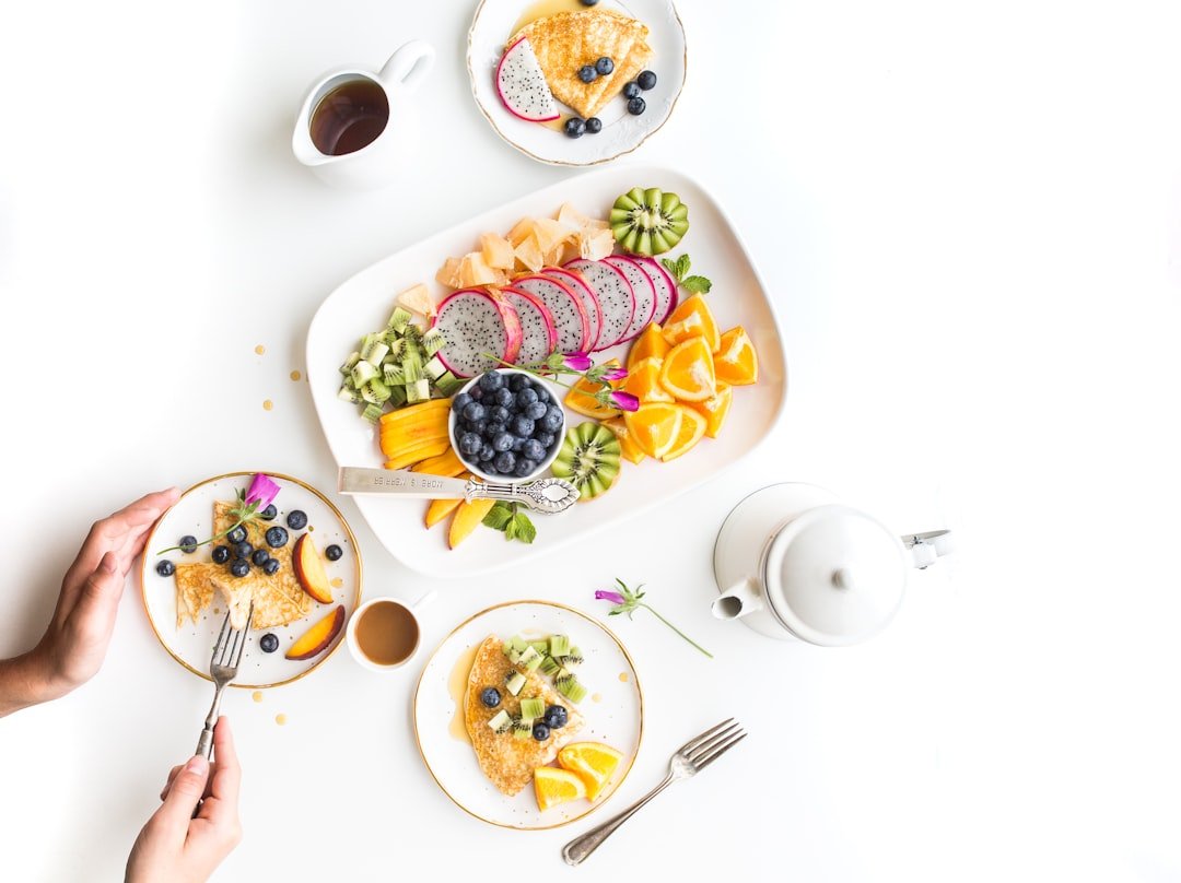 Der Food Fotograf - Eine Person isst eine Mahlzeit mit zwei Tellern Pfannkuchen, belegt mit Kiwi, Blaubeeren und Orangenscheiben. Auf einer großen Platte in der Mitte liegen verschiedene Früchte, darunter Drachenfrucht, Kiwi und Orangen. Auf dem Tisch stehen auch eine Teekanne, eine Tasse und eine Untertasse. Lecker Essen fotografieren.