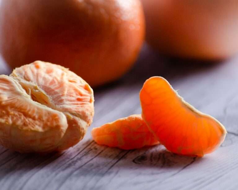 Food Fotograf Hannover - Nahaufnahme einer geschälten Mandarine mit drei abgetrennten Segmenten, die auf einer hellen Holzoberfläche liegen. Eine weitere ganze Mandarine ist im Hintergrund verschwommen. Der Fokus liegt auf der leuchtend orangen Farbe und Textur der Fruchtsegmente. Lecker Essen fotografieren.