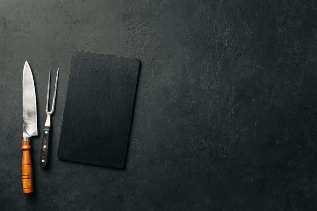Food Fotograf Hannover - Ein großes Küchenmesser mit Holzgriff, eine Fleischgabel und ein rechteckiges schwarzes Schneidebrett liegen in einer Reihe auf einer dunklen Steinoberfläche. Diese elegante Anordnung ist ein idealer Hintergrund für die Lebensmittelfotografie, wobei die Gegenstände links platziert sind und rechts viel leerer Platz bleibt. Food Fotografie Hannover.