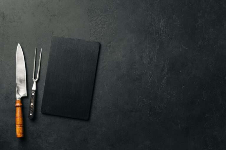 Food Fotograf Hannover - Ein großes Küchenmesser mit Holzgriff, eine Fleischgabel und ein rechteckiges schwarzes Schneidebrett liegen in einer Reihe auf einer dunklen Steinoberfläche. Diese elegante Anordnung ist ein idealer Hintergrund für die Lebensmittelfotografie, wobei die Gegenstände links platziert sind und rechts viel leerer Platz bleibt. Food Fotografie Hannover.