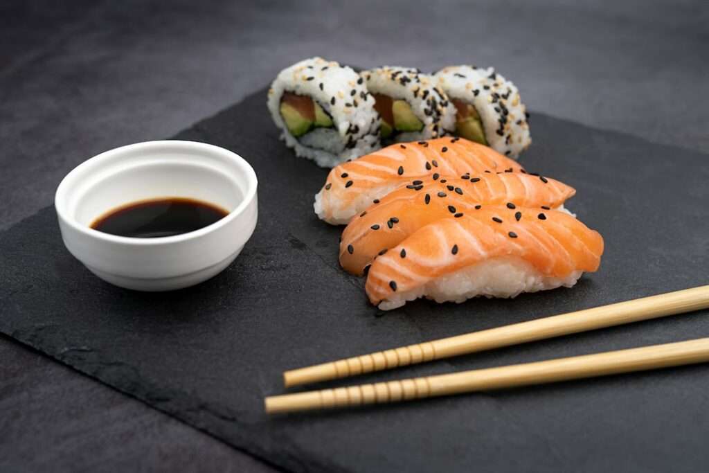 Food Fotograf Hannover - Ein Teller Sushi mit Lachs-Nigiri-Scheiben, garniert mit schwarzem Sesam, und mit Avocado und Thunfisch gefüllten Maki-Rollen, serviert mit einer kleinen Schüssel Sojasauce und einem Paar Holzstäbchen auf einer dunklen Schieferplatte. Food Fotografie Hannover.