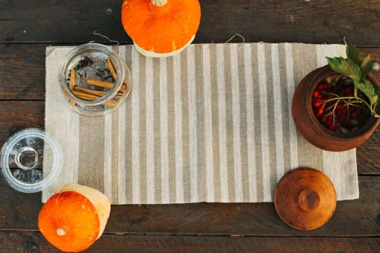 Food Fotograf Hannover - Eine rustikale Tischdekoration, die sich ideal als Hintergrund für Food-Fotografie eignet, besteht aus einem beige-weiß gestreiften Tischläufer, zwei kleinen orange-weißen Kürbissen, einem Glasgefäß mit Gewürzstäbchen, einem Holzbehälter mit roten Beeren und Blättern und einem geschlossenen Holzdeckel auf einem Holztisch. Food Fotografie Hannover.