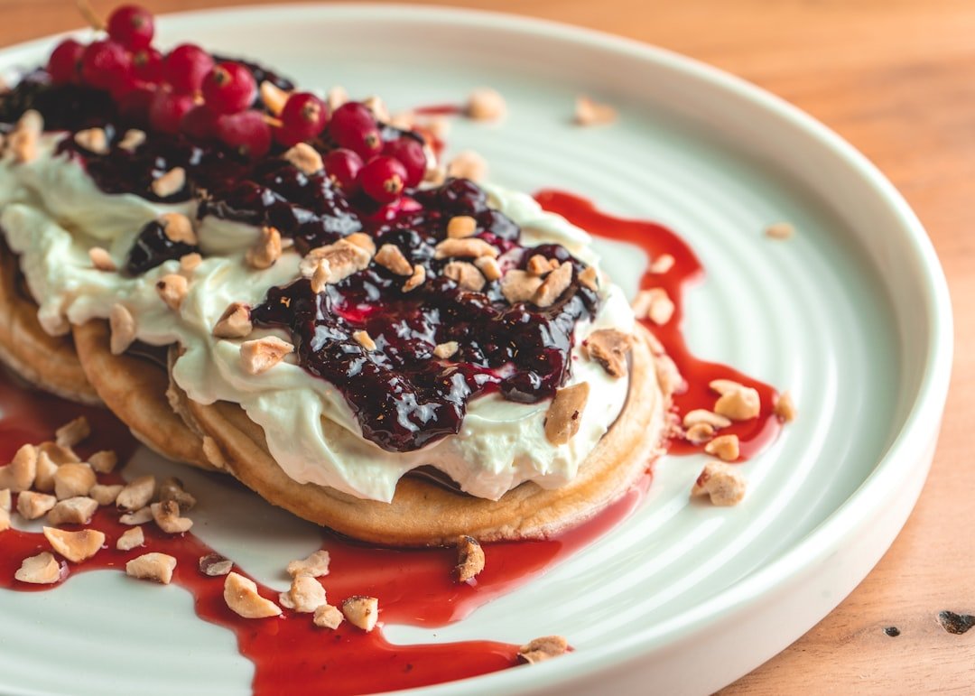 Der Food Fotograf - Auf einem Teller liegt ein Dessert mit Gebäck, das mit Schlagsahne und dunkler Beerensauce belegt und mit gehackten Nüssen bestreut ist. Obenauf liegen rote Beeren an einem Stiel. Das Gericht steht auf einem weißen Teller auf einer Holzoberfläche. Lecker Essen fotografieren.