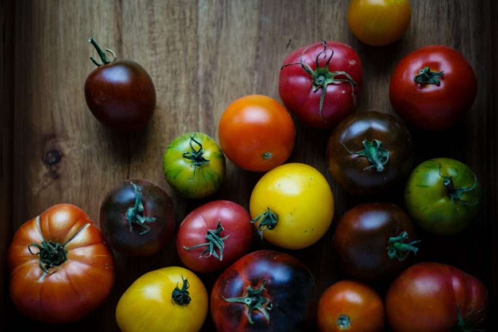 Food Fotograf Hannover - Eine Holzoberfläche ist mit verschiedenen Sorten von Heirloom-Tomaten in verschiedenen Farben wie Rot, Gelb, Grün und Braun bedeckt. Einige Tomaten haben Stiele und sind unterschiedlich groß. Die Beleuchtung betont die natürlichen Farben und Texturen der Tomaten. Food Fotografie Hannover.
