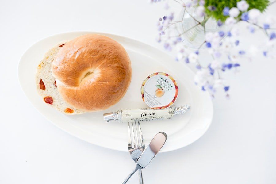 Der Food Fotograf - Ein Teller mit einem halbierten Bagel, einem kleinen Behälter mit Aprikosenmarmelade und einem eingewickelten Stück Käse. Auf dem Tellerrand liegen eine Gabel und ein Messer, und auf der rechten Seite ist eine verschwommene Vase mit violetten und weißen Blumen zu sehen. Lecker Essen fotografieren.