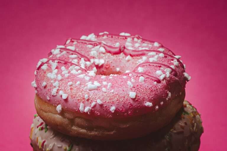 Food Fotograf Hannover - Eine Nahaufnahme eines Donuts mit rosa Zuckerguss, weißen Streuseln und einem Spritzer rosa Zuckerguss obendrauf, vor einem rosa Hintergrund. Nur die obere Hälfte eines anderen Donuts ist unten im Bild zu sehen. Food Fotografie Hannover.