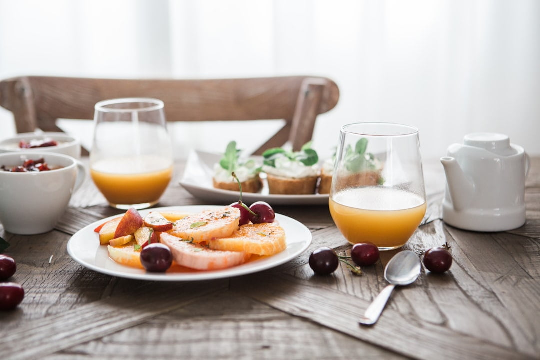 Der Food Fotograf - Ein Holztisch mit verschiedenen Frühstücksgerichten. Es gibt zwei Gläser Orangensaft, einen weißen Teller mit Grapefruitscheiben, Apfelscheiben und Kirschen, zwei Schüsseln mit Belag, eine weiße Teekanne und einen kleinen Teller mit Gebäck, garniert mit Gemüse. Lecker Essen fotografieren.