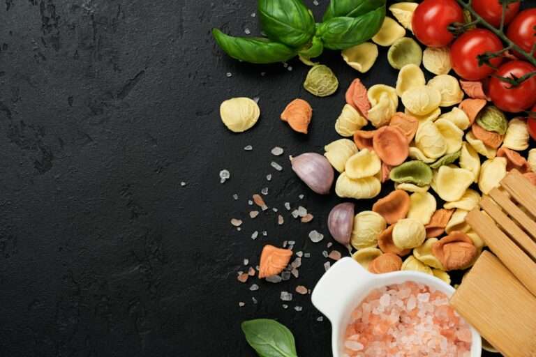 Food Fotograf Hannover - Eine Auswahl trockener, bunter Tortellini, Kirschtomaten, Knoblauchzehen, frische Basilikumblätter und ein Holzlöffel sind auf einer dunklen strukturierten Oberfläche ausgebreitet. Außerdem ist eine weiße Schüssel mit rosa Himalaya-Salz zu sehen. Food Fotografie Hannover.