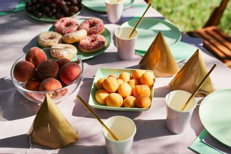 Food Fotograf Hannover - Ein für eine Party gedeckter Tisch. Es gibt Schalen mit Pfirsichen, Teller mit Donuts, Pappbecher mit Strohhalmen und goldene Partyhüte. Der Tisch ist mit einer hellen Tischdecke bedeckt und um ihn herum sind grüne Teller platziert. Die Lebensmittelfotografie fängt die lebendigen Farben perfekt ein, wobei Trauben den Hintergrund hervorheben. Food Fotografie Hannover.