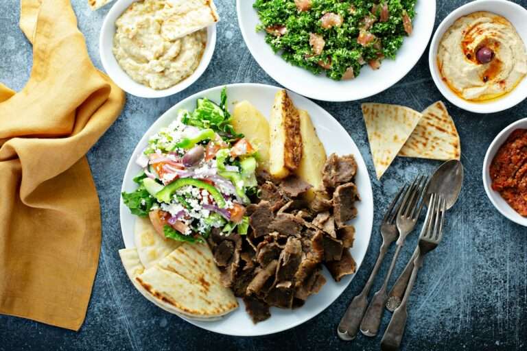 Food Fotograf Hannover - Ein Teller mit mediterranem Essen umfasst Gyrosfleisch vom Lamm, einen griechischen Salat mit Feta, Pita-Brot, Kartoffelecken und Dip-Saucen. Um den Teller herum stehen weitere kleine Schälchen mit mehr Salat, Brot und Saucen. Das Besteck ist rechts vor einem geschmackvollen Food-Fotografie-Hintergrund platziert. Food Fotografie Hannover.