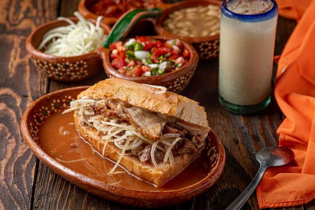 Food Fotograf Hannover - Ein Sandwich mit Fleisch und zerkleinertem Kohl liegt auf einem braunen Keramikteller. Auf dem Tisch, aufgenommen von Food Fotograf Hannover, stehen kleine Schälchen mit zerkleinertem Kohl, Salsa, Bohnen und ein Glas cremiges Getränk. Neben dem Teller auf der rustikalen Holzoberfläche stehen ein orangefarbenes Tuch und ein Löffel. Lecker Essen fotografieren.