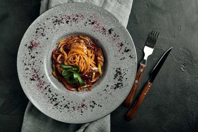 Food Fotograf Hannover - Ein Nudelgericht mit Spaghetti, Fleisch und Garnitur auf einem strukturierten, grauen Teller steht auf einem grauen Tuch. Eine Gabel und ein Messer mit Holzgriffen liegen rechts neben dem Teller auf einer dunklen, strukturierten Oberfläche. Food Fotografie Hannover.