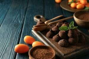 Food Fotograf Hannover - Auf einem Holzbrett werden mit Kakaopulver bestäubte Schokoladentrüffel präsentiert, elegant garniert mit Minzblättern. Daneben stehen frische Kumquats neben einer Schale Kakaopulver. Die Szene wird durch die richtige Beleuchtung in der Food-Fotografie vor einer dunkelblauen Holzoberfläche noch verstärkt. Food Fotografie Hannover.