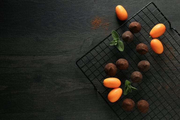 Food Fotograf Hannover - Auf einem Abkühlgitter sind Pralinen und Kumquats ordentlich angeordnet. Ein paar frische Minzblätter sind dazwischen auf einem dunklen, strukturierten Hintergrund für Lebensmittelfotografie verstreut, daneben liegt eine Prise Kakaopulver. Food Fotografie Hannover.