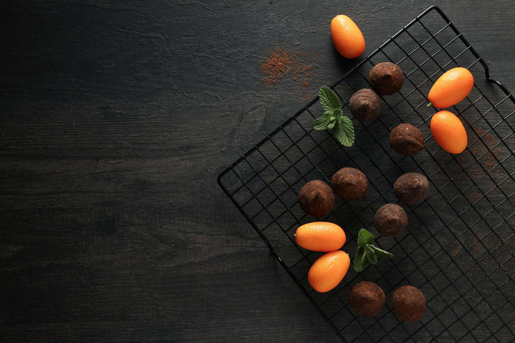 Food Fotograf Hannover - Auf einem Abkühlgitter sind Pralinen und Kumquats ordentlich angeordnet. Ein paar frische Minzblätter sind dazwischen auf einem dunklen, strukturierten Hintergrund für Lebensmittelfotografie verstreut, daneben liegt eine Prise Kakaopulver. Food Fotografie Hannover.