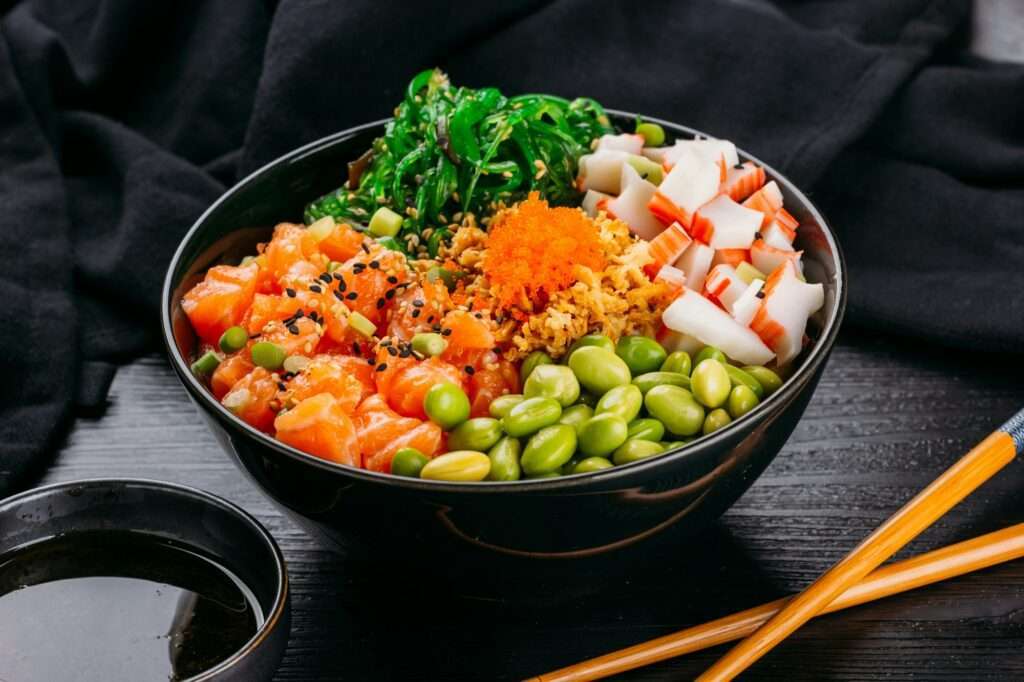 Food Fotograf Hannover - Eine Schüssel mit Sushi-Zutaten enthält gewürfelten Lachs, Edamame, Seetangsalat, Krabbenstäbchen, Sesamsamen und orangefarbenen Fischrogen. Ein kleiner Teller mit Sojasauce und ein Paar Essstäbchen stehen neben der Schüssel auf einer schwarzen Oberfläche. Food Fotografie Hannover.