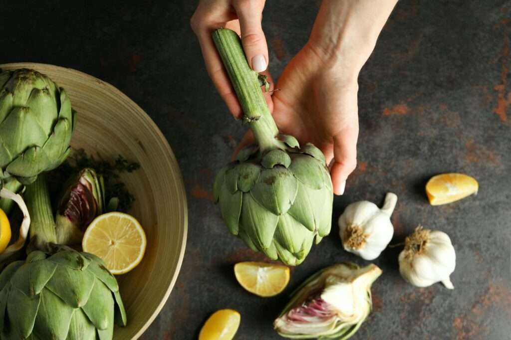 Food Fotograf Hannover - Eine Person hält eine Artischocke über einem dunklen Food-Fotografie-Hintergrund. Daneben steht eine Holzschale mit weiteren Artischocken, einer halben Zitrone, Knoblauchknollen und Zitronenscheiben. Eine weitere halbierte Artischocke liegt neben dem Knoblauch und schafft ein einladendes kulinarisches Tableau. Food Fotografie Hannover.