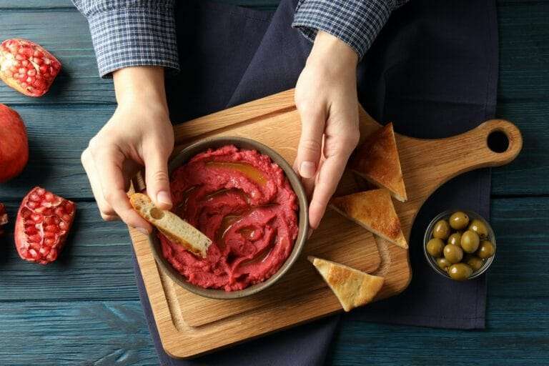 Food Fotograf Hannover - Eine Person taucht ein Stück Brot in eine Schüssel mit Rote-Bete-Hummus auf einem rustikalen Holzbrett und perfektioniert damit ihr Food-Fotografie-Setup. Um die Szene herum sind Pita-Dreiecke, eine kleine Schale mit grünen Oliven und halbierte Granatäpfel, alles auf einem dunklen Holztisch präsentiert. Food Fotografie Hannover.