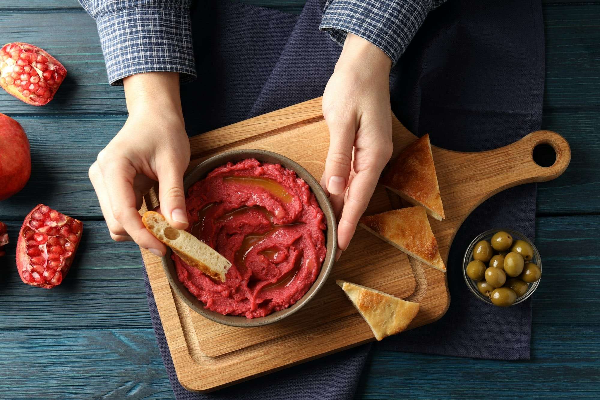Food Fotograf Hannover - Eine Person taucht ein Stück Brot in eine Schüssel mit Rote-Bete-Hummus auf einem rustikalen Holzbrett und perfektioniert damit ihr Food-Fotografie-Setup. Um die Szene herum sind Pita-Dreiecke, eine kleine Schale mit grünen Oliven und halbierte Granatäpfel, alles auf einem dunklen Holztisch präsentiert. Food Fotografie Hannover.