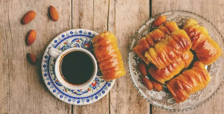 Food Fotograf Hannover - Eine Tasse schwarzen Kaffee steht auf einer dekorativen Untertasse und fängt die Essenz der Food-Fotografie ein. Daneben verführt ein Glasteller mit glänzendem, goldbraunem Gebäck die Sinne. Auf dem Holztisch im Hintergrund sind kunstvoll Mandeln verstreut. Food Fotografie Hannover.