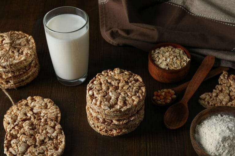 Food Fotograf Hannover - Ein Holztisch mit Reiskuchen, einem Glas Milch, kleinen Schüsseln mit Haferflocken und Maiskörnern und einem Holzlöffel mit Mehl. Im Hintergrund ist teilweise ein gefaltetes braunes Tuch zu sehen. Food Fotografie Hannover.