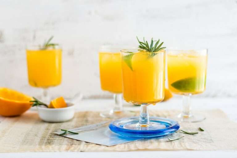 Food Fotograf Hannover - Vier Gläser Orangensaft mit Limettenscheiben und Rosmarinzweigen stehen auf einem Tisch. Ein Glas steht auf einem blauen Untersetzer. Darüber steht eine kleine weiße Schale mit Orangenscheiben und Rosmarinzweigen. Der Hintergrund ist eine helle, strukturierte Wand. Food Fotografie Hannover.