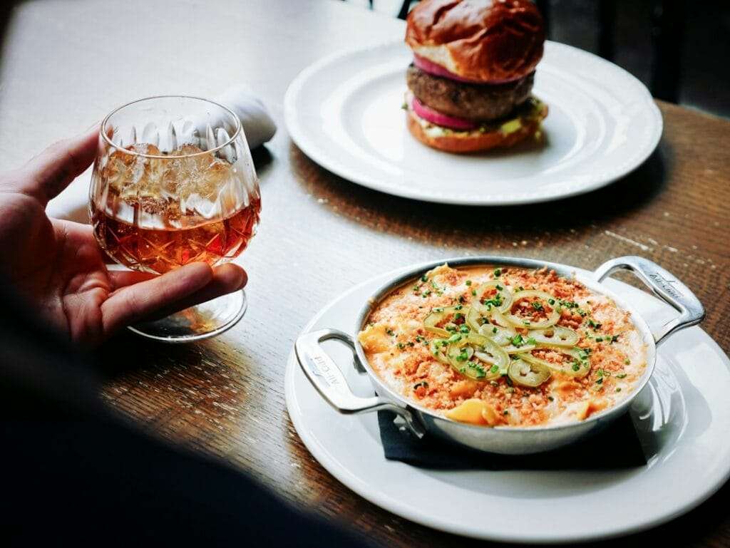 Food Fotograf Hannover - Die Hand einer Person hält vorsichtig ein Glas Whiskey on the Rocks und fängt einen Moment kulinarischer Glückseligkeit ein. In dieser Food-Fotografie-Szene lugt ein Burger aus dem Hintergrund hervor, während gebackene Makkaroni mit Käse und Frühlingszwiebeln den Vordergrund einnehmen. Food Fotografie Hannover.