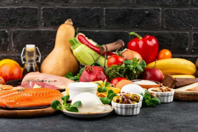 Food Fotograf Hannover - In dieser eindrucksvollen Food-Fotografie werden vor einem dunklen Hintergrund kunstvoll verschiedene Lebensmittel präsentiert. Roher Lachs, Mozzarella, Eier und frisches Gemüse mischen sich mit Früchten, Nüssen und Brot. Eine Flasche Öl verleiht der Komposition einen Hauch von Flair, und eine Backsteinmauer sorgt für einen stimmungsvollen Hintergrund. Food Fotografie Hannover.