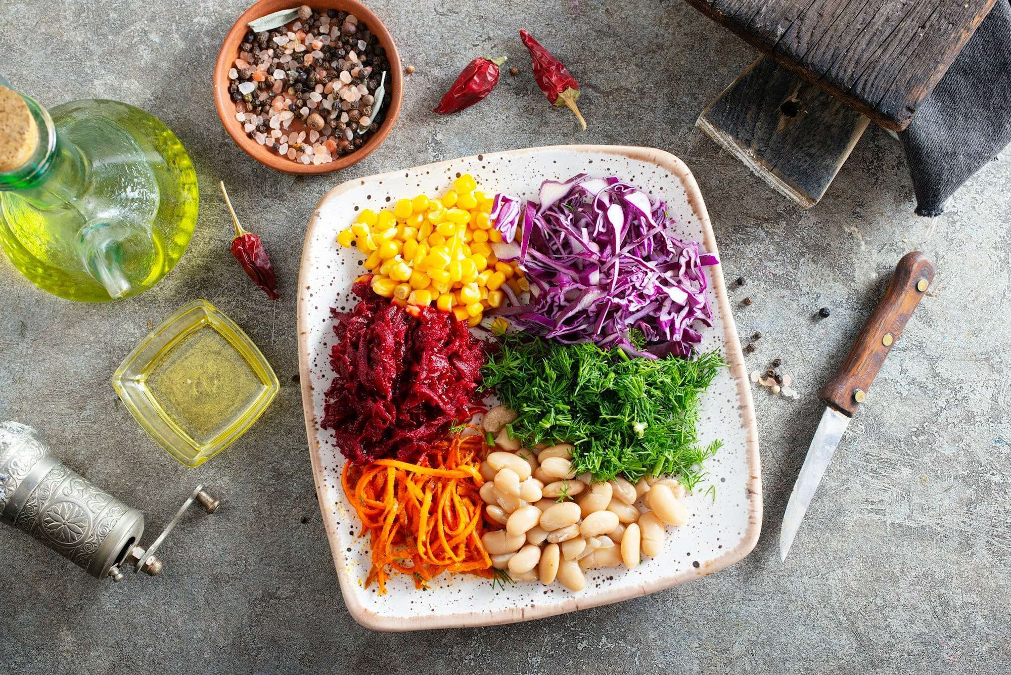 Food Fotograf Hannover - Ein Teller mit verschiedenen Gemüsesorten, darunter Mais, zerkleinerter Rotkohl, geriebene Rote Bete, geriebene Karotten, Dill und weiße Bohnen, steht auf einer strukturierten Oberfläche – perfekt für Food-Fotografie. In der Nähe befinden sich ein Messer, eine Schüssel mit rosa Steinsalz, Chilischoten und eine Flasche Olivenöl. Food Fotografie Hannover.