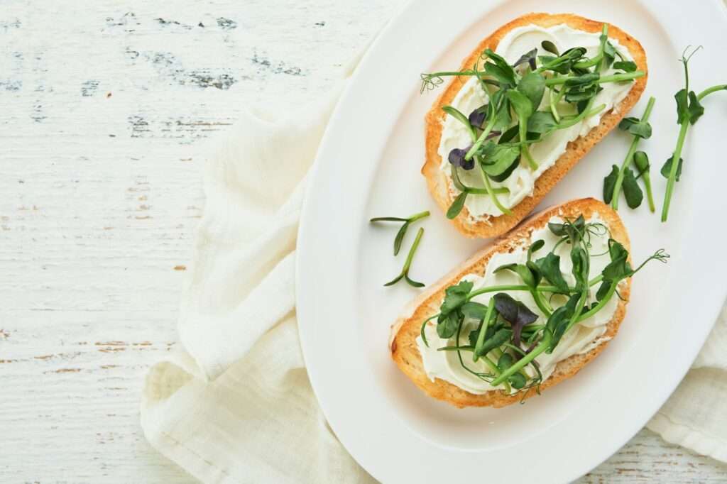 Food Fotograf Hannover - Zwei Brotscheiben mit Frischkäse, Gurkenscheiben und einer Mischung aus Microgreens sind kunstvoll auf einem weißen ovalen Teller arrangiert. Die Szene ist vor einem hellen Stoffhintergrund inszeniert und fängt die Essenz der Food-Fotografie auf einer weißen Holzoberfläche ein. Food Fotografie Hannover.