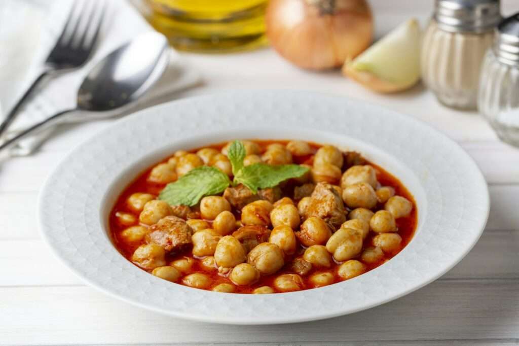 Food Fotograf Hannover - Ein weißer Teller mit Kichererbseneintopf, garniert mit einem frischen Minzblatt, sitzt elegant in der Food-Fotografie. Der Eintopf enthält Kichererbsen und Fleischstücke in einer Tomatensauce. Im Hintergrund befinden sich eine Zwiebel, eine Knoblauchzehe, Öl sowie Salz- und Pfefferstreuer, die dem Hintergrund Tiefe verleihen. Food Fotografie Hannover.