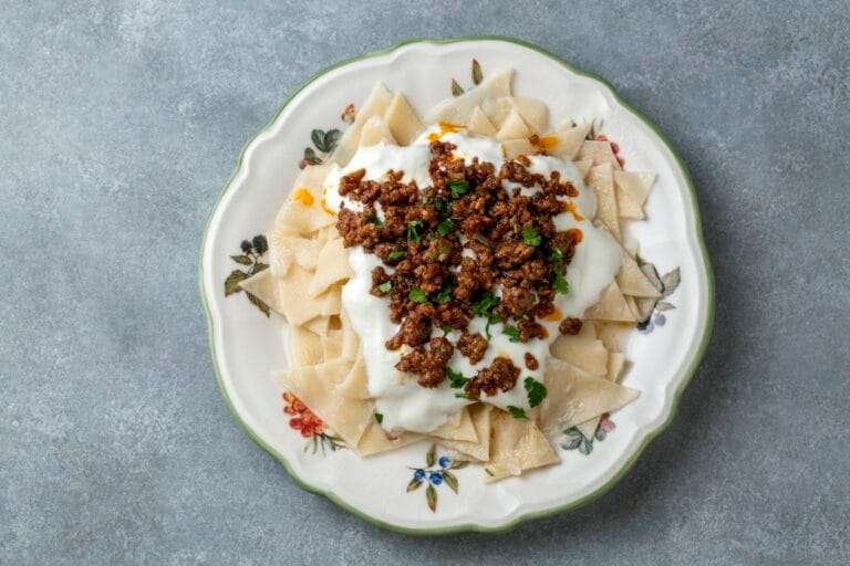 Food Fotograf Hannover - Ein Teller mit traditionellem türkischem Mantı, belegt mit Joghurt und gewürztem Hackfleisch, steht auf einem Teller mit Blumenmuster. Der Hintergrund ist eine strukturierte graue Oberfläche. Food Fotografie Hannover.