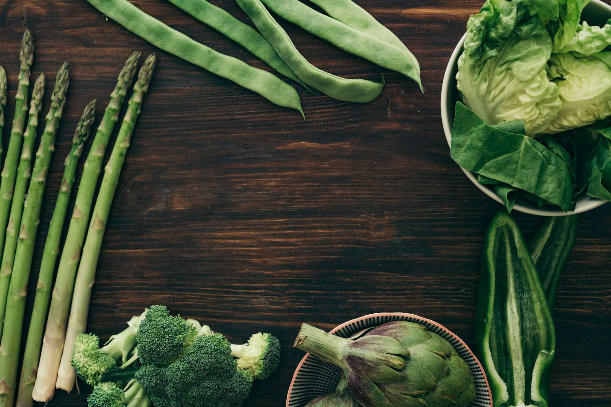 Food Fotograf Hannover - Auf einer Holzoberfläche sind verschiedene grüne Gemüsesorten angerichtet, darunter Spargel, grüne Bohnen, Brokkoli, eine Artischocke und Blattgemüse in einer Schüssel. Food Fotografie Hannover.