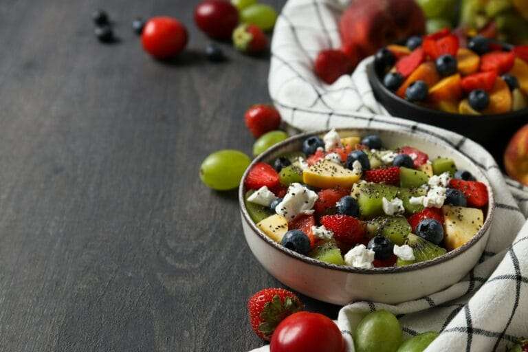 Food Fotograf Hannover - Eine Schüssel Obstsalat mit Erdbeeren, Blaubeeren, Kiwi, Trauben und Käsestreuseln steht auf einer dunklen Holzoberfläche – perfekt für jede Art von Food-Fotografie. Im Hintergrund halten ein weißes Tuch und eine weitere Schüssel eine ähnliche Mischung vor einem einladenden Hintergrund aus verstreuten Früchten. Food Fotografie Hannover.