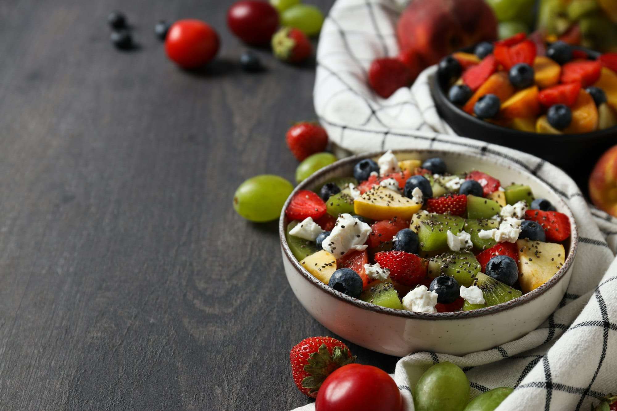 Food Fotograf Hannover - Eine Schüssel Obstsalat mit Erdbeeren, Blaubeeren, Kiwi, Trauben und Käsestreuseln steht auf einer dunklen Holzoberfläche – perfekt für jede Art von Food-Fotografie. Im Hintergrund halten ein weißes Tuch und eine weitere Schüssel eine ähnliche Mischung vor einem einladenden Hintergrund aus verstreuten Früchten. Food Fotografie Hannover.